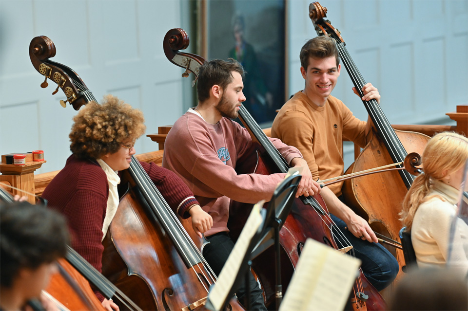 A group of students, with their double bass next to them, smiling and chatting in a orchestra performance.
